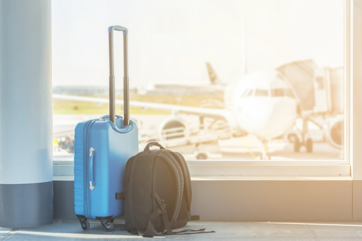 En koffert og en sekk står foran et vindu på en flyplass. Utenfor vinduet står et fly. Bildet illustrerer en flytteprosess til et annet land.